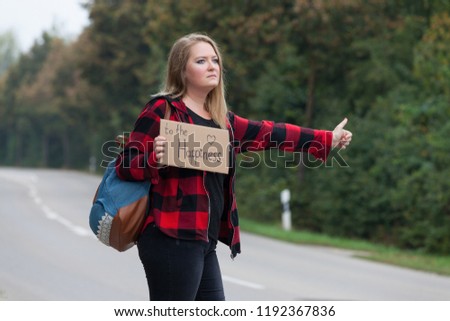 A young woman in a checkered shirt and with a backpack is hitchhiking with a cardboard sign in her hands.