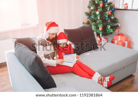 Merry Christmas and Happy New Year. Young woman sit on sofa with daughter. She holds book with pictures on her lap. Woman is reading. Girl listen. They look calm ad peaceful.