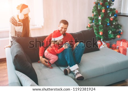 Merry Christmas and Happy New Year. Family picture. Young man sits on sofa with daughter. They spend time tgether. Woman stands at sofa. She looks at them. Kid reaches and touches her dad's knee.