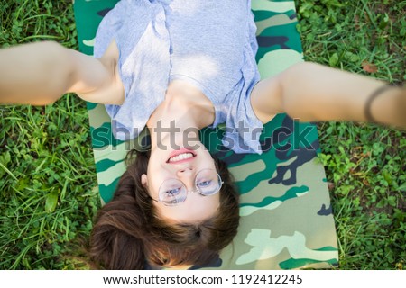 Close up of laughing brunette woman in eyeglasses lying on grass in park and making selfie
