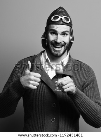 handsome bearded aviator man with long beard on happy face holding glass of alcoholic shot in gray knitted sweater with hat and glasses on blue studio background.