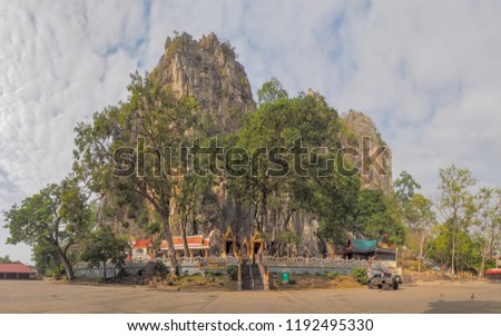 view of Wat Tham Khao Yoi Cave with cloudy sky background, Buddhist temple in the rock mountain, Phetchaburi, Thailand with Thai text banner means the name of this local is Wat Tham Chao Yoi Cave.