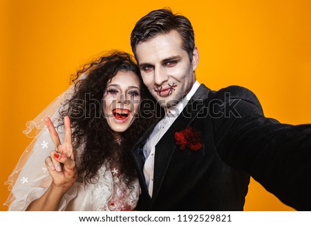 Photo of beautiful zombie couple bridegroom and bride wearing wedding outfit and halloween makeup laughing while taking selfie isolated over yellow background