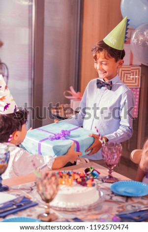 Present for boy. Cheerful kind boy smiling and wearing party hat while giving present to his friend