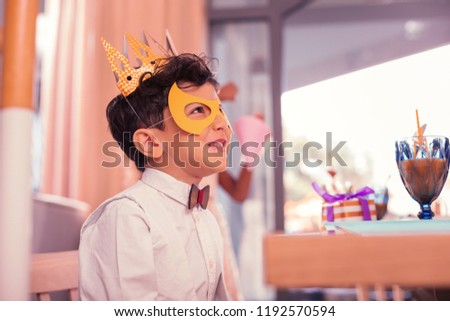 Boy wearing mask. Emotional dark haired kid smiling and looking into the distance while wearing paper crown and mask at the carnival