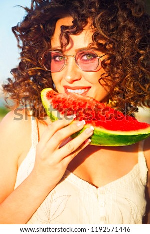 Beauty smiling curly woman is wearing pink sunglasses and eating watermelon on the beach