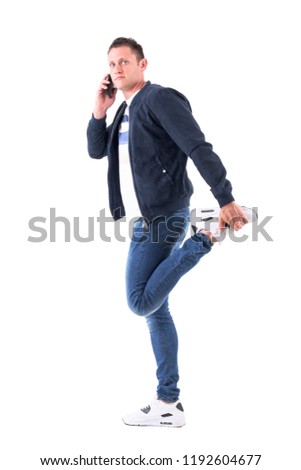 Young adult man in bomber jacket talking on the phone and stretching leg. Full body isolated on white background.