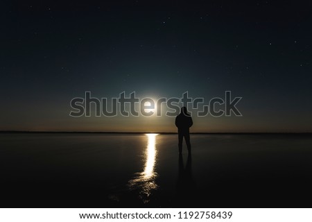 The man stands at the sunset of the moon under the starry sky with bright stars and milk way. Silhouetted photo against the starry night sky. Reflection of the rays from the moon in the water.