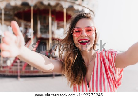Lovely female model in pink outfit making selfie in amusement park. Stunning elegant young woman enjoying good summer day.