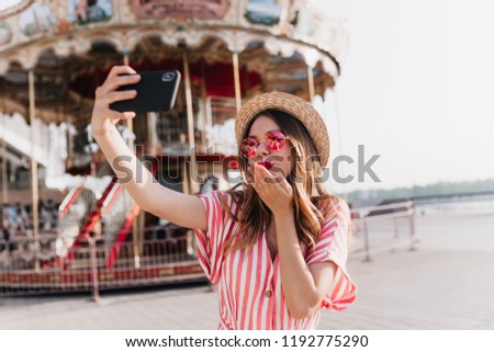 Inspired caucasian girl in straw hat sending air kiss for selfie. Outdoor shot of fascinating woman in pink sunglasses taking picture of herself near carousel.