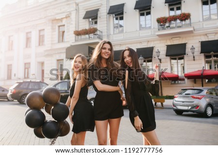 Outdoor portrait of slim girls having fun together after party and walking down the street. Stunning young female models posing with helium balloons in front of cars and beautiful old building.