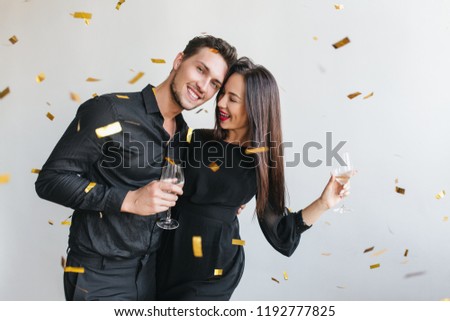 Glad european man embracing his wife at friend's birthday party. Young couple in black attire posing with champagne at light background under confetti.