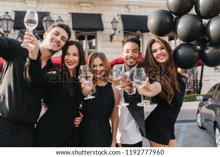 Graceful girl with long light-brown hair holding black helium balloons in front of building and laughing. Cute blonde woman raising glass of champagne while posing with african friend.