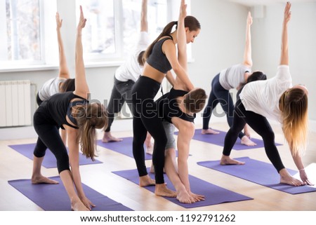 Female yoga instructor teaching Utthita Trikonasana pose, extended triangle exercise for a group of sporty people practicing in studio, working out indoor, teacher helping to master, full length Royalty-Free Stock Photo #1192791262