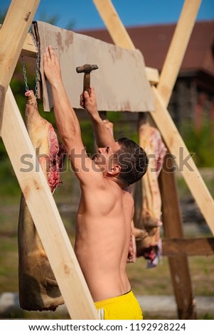 Young Caucasian guy without a shirt pins a plate to the frame with pork carcasses