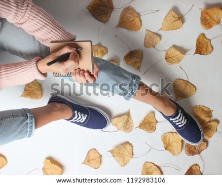 A girl in jeans and sneakers sits on  white background among the fallen yellow leaves and writes in a notebook. Work journalist. Autumn time. Working space.
