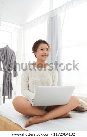 Pretty smiling young woman sitting on bed and working on laptop