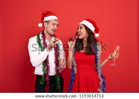Portrait young couple holding glasses of champagne and  blowing party whistles celebrating on red background. christmas and happy new year concept. 
