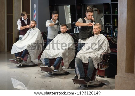 Process of styling and grooming men's haircuts in barbershop. Three professional and confident barbers standing and cutting hair of men. Male clients sitting in chairs and wearing haircut gowns. Royalty-Free Stock Photo #1193120149