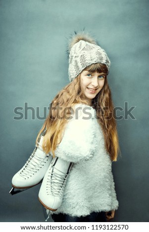 A girl with skates in her hands in a white sweater and hat.