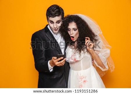 Surprised creepy man and woman in halloween wedding costumes using smartphone isolated over orange