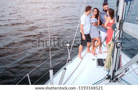 Nice picture of four people standing on yacht. Brunette is looking at guy. She smiles. He embraces her. Another couple standing close too. Man with beard smiles too.