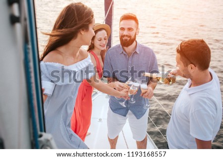 Nice picture of four people standing together on yacht. Man in white shirt pours champaigne into glasses. Brunette looks at another man. He smiles. There is another woman behind.
