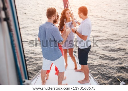 Picture of three people looking at brunette. They talk to her. She smiles and look at them. People stand at bow of yacht. Sun goes down.