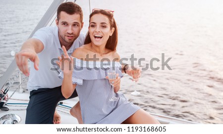 Nice picture of couple sitting together very close. Guy points forward. Woman is excited. She points up and smile a bit. They hold glasses of champaigne.