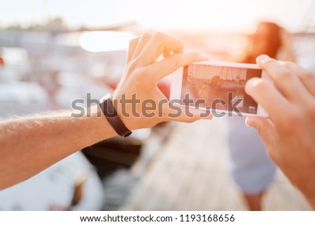 Man takes picture of young woman. She poses on camera and smiles. Brunette stands on pier. There are yachts on each side of it.