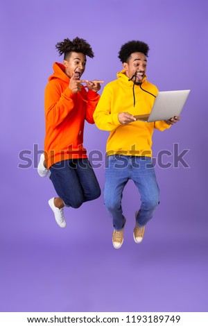 Full length of two happy male african friends in colorful hoodies jumping isolated over violet background, holding laptop computer