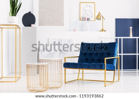 Two stylish tables next to petrol blue armchair in bright living room interior of modern apartment, real photo