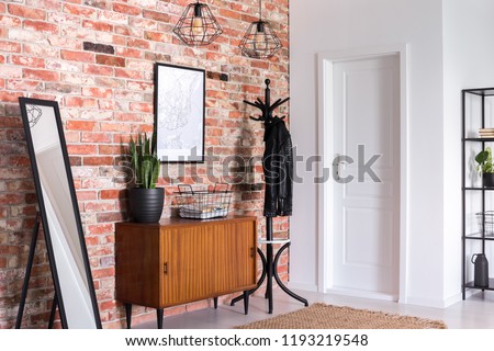 Mirror next to wooden cabinet in entrance hall interior with white door and poster on red brick wall. Real photo Royalty-Free Stock Photo #1193219548