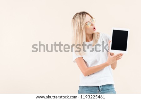 surprised stylish woman in eyeglasses showing digital tablet with blank screen isolated on beige