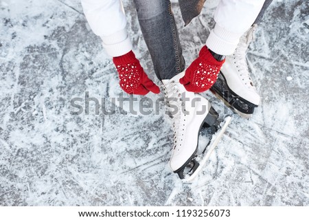 Girl tying shoelaces on ice skates before skating on the ice rink, hands in red knitted gloves. View from top. Royalty-Free Stock Photo #1193256073