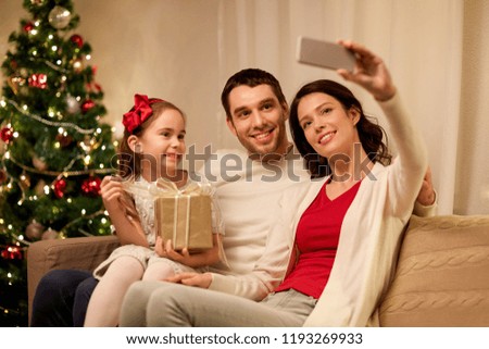 holidays, family and technology concept - happy mother, father and little daughter with christmas gift taking selfie by smartphone at home