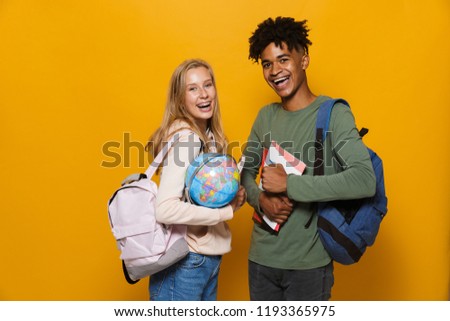 Photo of american and caucasian students man and woman 16-18 wearing backpacks holding earth globe isolated over yellow background