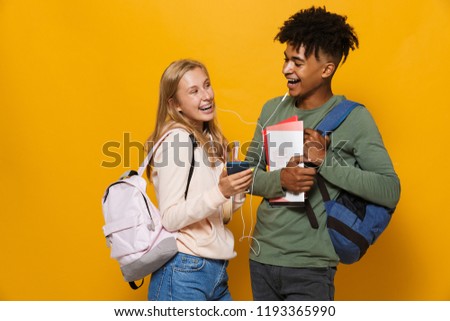 Photo of cheerful students man and woman 16-18 wearing earphones using mobile phones and holding exercise books isolated over yellow background