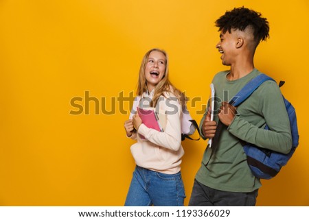Photo of positive people guy and girl 16-18 wearing backpacks smiling and holding exercise books while walking isolated over yellow background