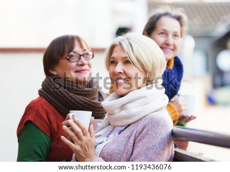 Happy senior woman in warm clothes having cup of hot tea on terrace. Focus on blonde woman