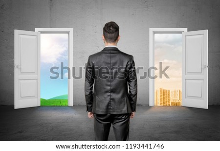 A back view on a businessman choosing between two doors, with a green field and with an apartment block. Construction and environment. City or village life.