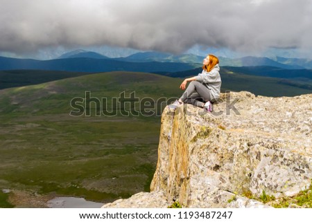 The beautiful red-haired woman sits and relaxing keep calm alone on the edge of a cliff in the rays of light on a rock Altai mountains on a background of blue sky with big gray clouds and landscape