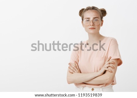 Studio shot of smart confident attractive european woman with cute freckles and buns hairstyle, wearing glasses and pink t-shirt, holding hands crossed on chest, gazing self-assured at camera