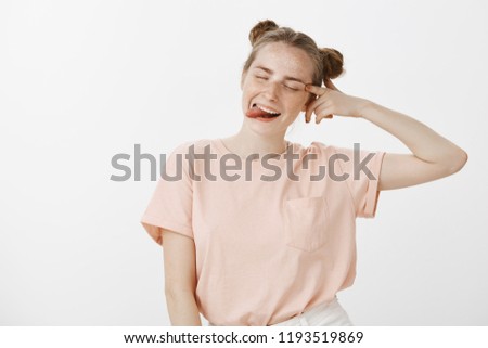 Waist-up shot of carefree joyful stylish european woman with buns hairstyle, closing eyes and touching eye with finger, sticking out tongue playfully, smiling broadly over gray wall, feeling great