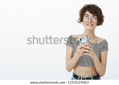 Music inspired girl to draw new picture. Stylish dreamy woman with short cute haircut and glasses, smiling broadly, gazing at upper left corner, listening songs in earphones and holding smartphone