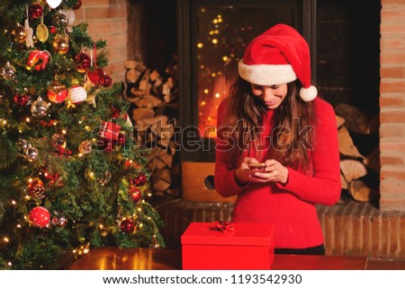 A beautiful woman at Christmas day sends messages, takes pictures and calls with her phone; in the background the Christmas tree and the lit fireplace. Concept of: christmas, promotions, technology.