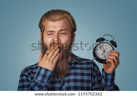 Businessman yawning with a alarm clock, being late, sleepless, sleepy man. Hipster man with beard in blue plaid checkered shirt  Isolated on blue Background. Negative face expression, human emotion.
