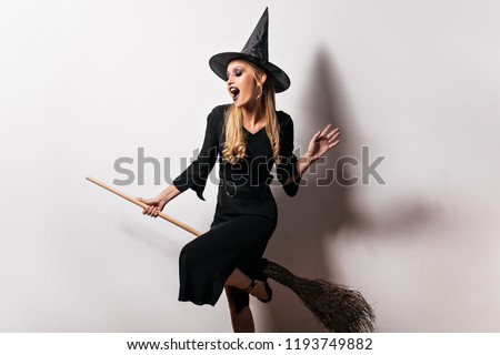 Joyful witch flying on broom in halloween. Indoor portrait of enthusiastic female wizard in black dress.