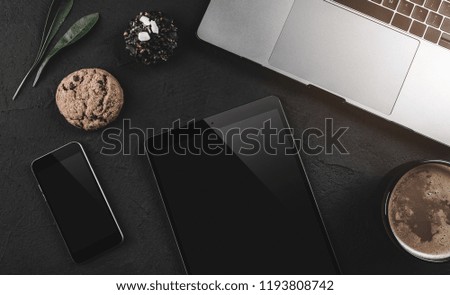 Top view of modern laptop, gadgets and delicious biscuits of chocolate on black stone background. Light effect in the top corner, flat lay