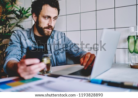 Pensive young man installing app on laptop and sharing media on smartphone using wireless internet connection.Concentrated male freelancer watching tutorial on website on netbook in cafe